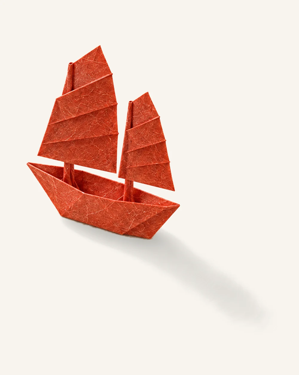 A small origami sailboat with two sails, folded from a single sheet of vermilion washi paper, on warm white paper.